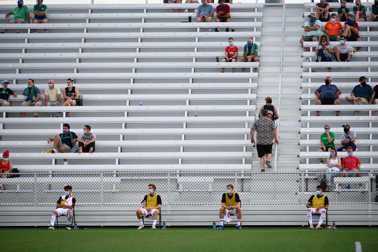 Members of Loudoun United FC's bench sit distanced apart wearing masks. 